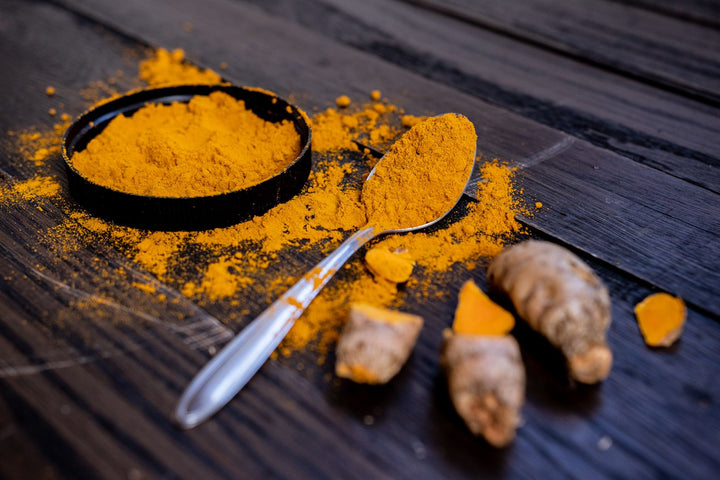 Ground turmeric in a small black dish with a spoon and fresh turmeric roots on a dark wooden surface