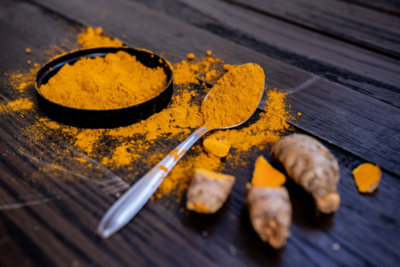 Ground turmeric in a small black dish with a spoon and fresh turmeric roots on a dark wooden surface
